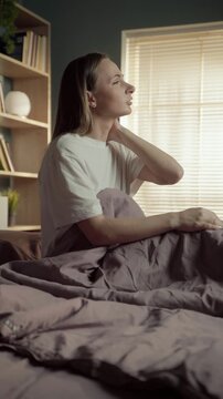 Young woman waking up in her bed and massaging her stiff neck, experiencing discomfort and muscle pain from sleeping in an awkward position or on an uncomfortable mattress and pillow