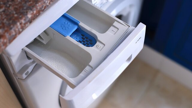 Washing machine detergent drawer with blue compartment for liquid detergent and white section for powder, positioned in a modern laundry room setting