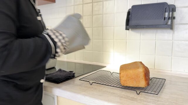 A person wearing oven mitts carefully places several loaves of freshly baked, hot bread onto a wire cooling rack in a kitchen. The process shows homemade baking and cooking from scratch