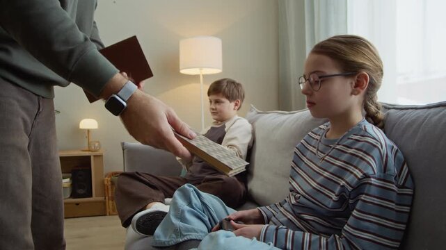 Handheld shot of father giving books to siblings and asking to read instead of playing video games with joysticks in living room