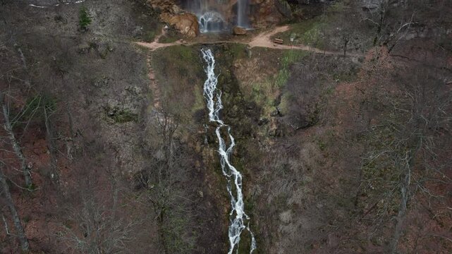 Aerial drone shot from top down perspective following a narrow waterfall stream down through forest terrain to its base, revealing hiking trail and natural surroundings in a scenic wilderness