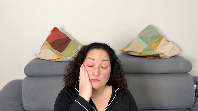 A middle aged woman with dark curly hair sits on a gray couch and uses a white electronic skincare device on her face