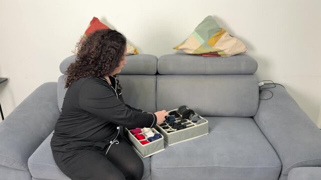 Woman in pajamas sits on a sofa, neatly folding socks into drawer organizers. Video shows process of tidying up and decluttering, ending with neatly arranged sock boxes. Home organization concept