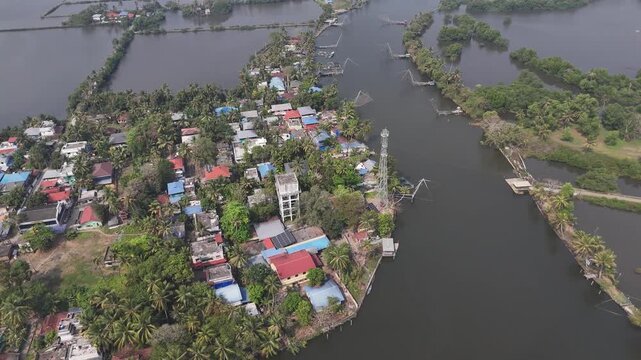 Drone footage captures a coastal Kerala village nestled among coconut palms along dark backwater channels. Colorful rooftops in red and blue dot the landscape while traditional Chinese fishing nets ex