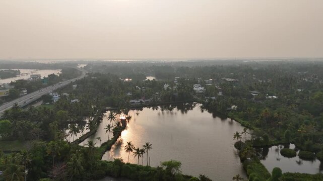 Golden sunset reflects on tranquil Kerala backwater lakes surrounded by lush coconut palm groves. Aerial drone footage reveals scattered houses and a highway bridge crossing the waterway in misty trop