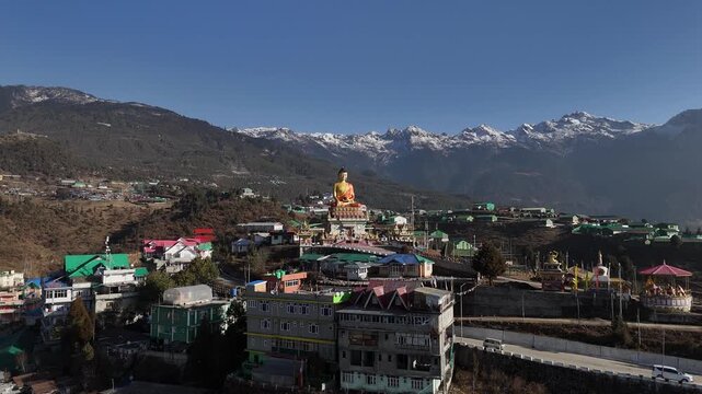 Drone footage captures the vibrant hillside town of Tawang featuring a golden Buddha statue overlooking colorful buildings with green and red rooftops against snow-capped Himalayan peaks under clear b