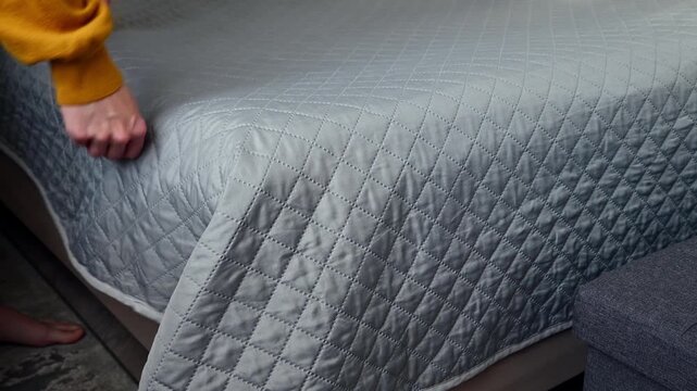 Person arranging bed linens, smoothing quilt on mattress in cozy bedroom setting