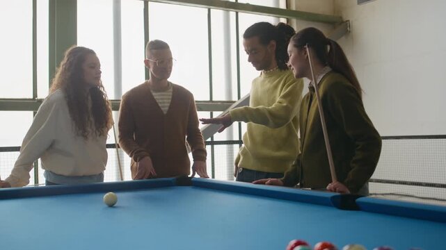 Handheld medium shot of young Caucasian woman making powerful break shot and hitting triangle rack while playing billiards with diverse friends in indoor setting