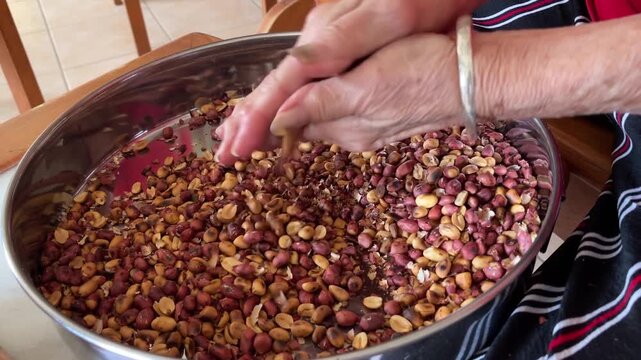 Hands rubbing and peeling skins from a large quantity of roasted peanuts in a metal bowl, preparing clean ingredients for traditional handmade cooking.