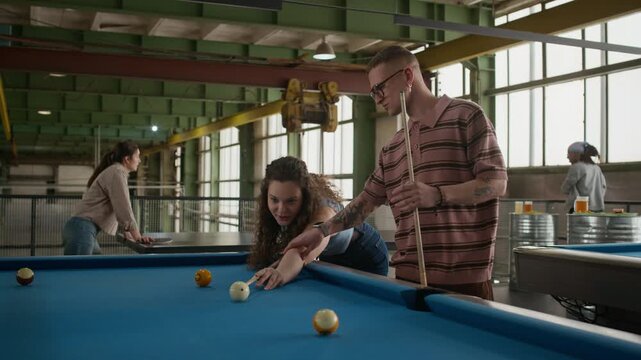 Medium shot of man guiding girlfriend with cue while teaching her to play billiards at blue table, another couple playing table tennis in background in industrial loft
