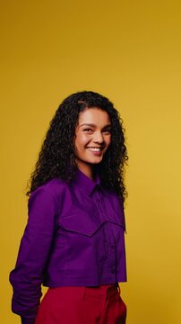 Young woman with curly hair smiles and plays with her hands on a yellow background in a vertical medium shot.