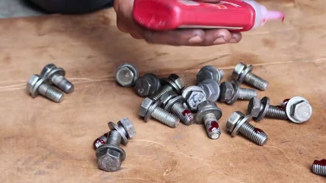 A technician uses a bottle to apply red high-strength threadlocker adhesive liquid to the threads of several metal flange bolts on a wooden workbench.