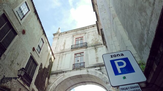 Low angle view revealing a beautiful stone archway between two weathered historic buildings in a narrow alley in Lisbon, Portugal, with a parking zone sign visible in the foreground. Arco Grande de Ci