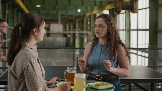 Medium shot of two Caucasian young women enjoying beer with snacks and conversation while two men playing ping pong in background, spending leisure time together in indoor activity club
