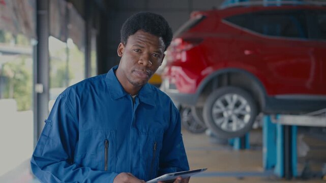 Young mechanic worker in blue uniform inside auto repair garage inspecting vehicle with tablet near red suv on lift, focused repair service and maintenance work in workshop over soft natural light