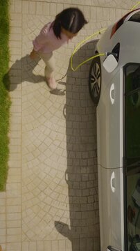 A woman walks toward a sleek gray electric car parked on a cobblestone driveway, carefully connecting a bright yellow charging cable to its port.