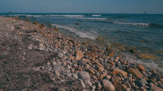 During midday, waves hit the rocky shore as the ocean glistens. With clear skies, the shore is lined with stones and the distant sight of boats can be seen.