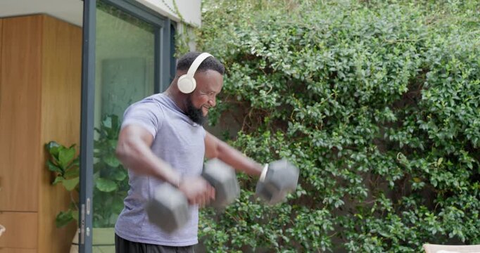 African American man in shorts stepping out from sliding door and training with dumbbells on patio
