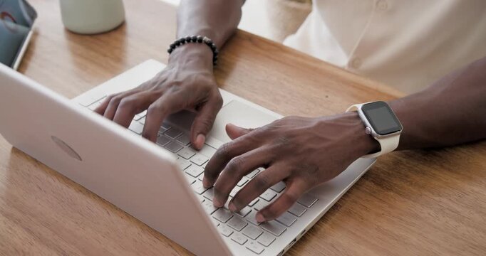 Adult African man placing hands, typing on laptop at desk, wearing smartwatch and bracelet, working
