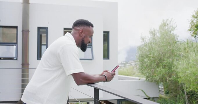 Receiving notification, adult African man leaning on balcony rail, scrolling red phone, responding