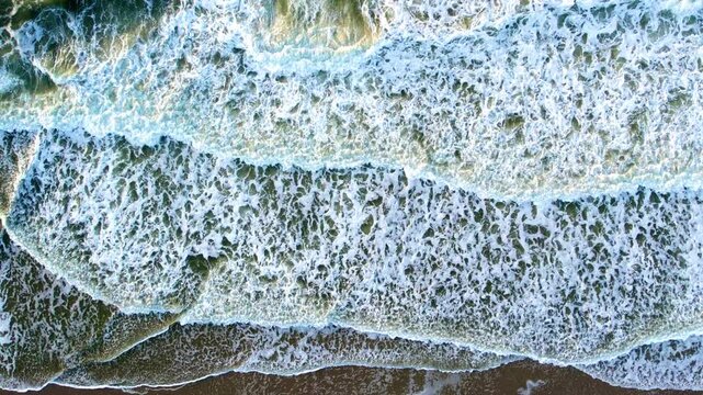 Aerial Top Down View of Ocean Waves Crashing on a Sandy Beach. Drone Footage of Sea Surf and White Foam.