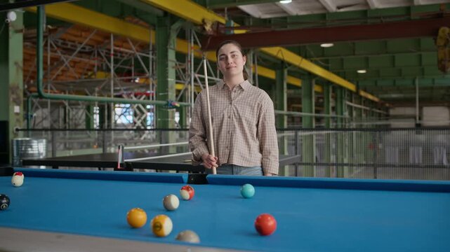 Medium portrait of young Caucasian woman holding billiard cue and posing confidently at blue pool table in industrial loft setting