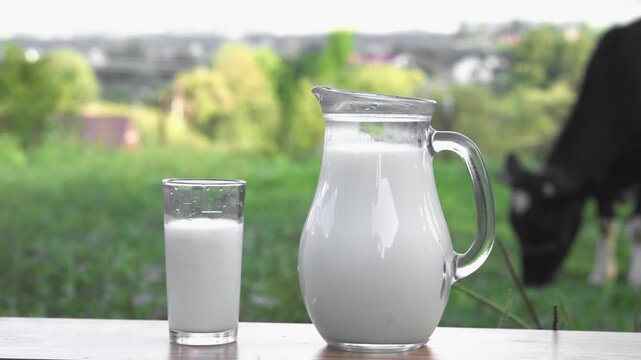 A glass and jug filled with fresh milk are placed on a wooden surface, set against a lush green field and a grazing cow. The scene highlights organic farming and healthy nutrition