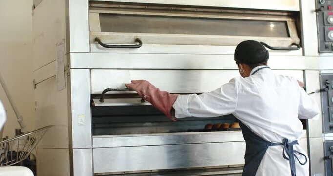 Vertical video: Opening oven, bakery baker in long mitts pulling tray of rolls to check, copy space