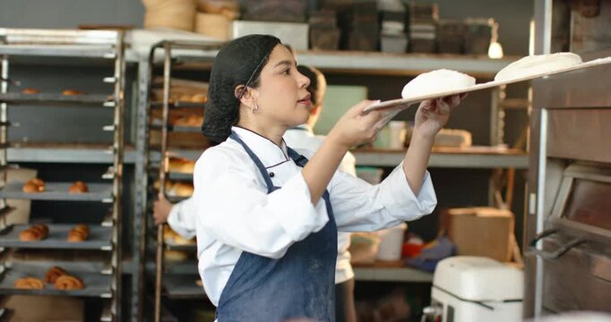 Diverse female team in white jackets holding floured tray sliding it into oven for baking bread