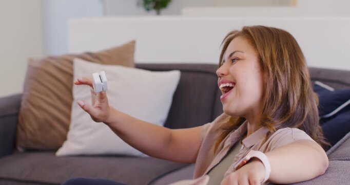 African American woman holding white device, examining, reacting to sound from device on gray couch