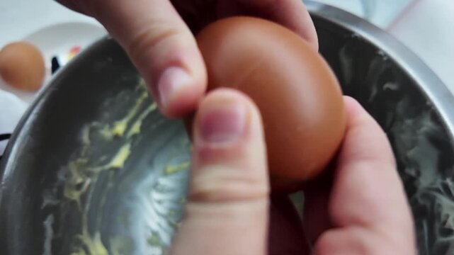 Slow motion close-up of a child's hands trying to crack an egg over a metal bowl. Shell breaks, yolk misses the bowl and spills over hands. Messy cooking moment.