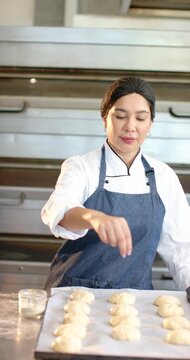 Mid-adult chef leaning, sprinkling topping on tray of dough at steel counter preparing for baking