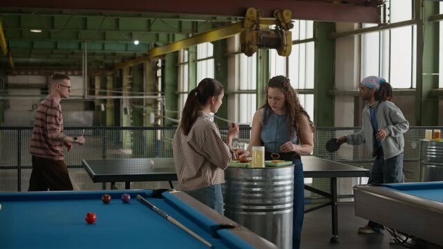 Medium wide shot of two Caucasian female friends standing by keg table sharing beer and conversation while men playing table tennis in background in spacious industrial loft