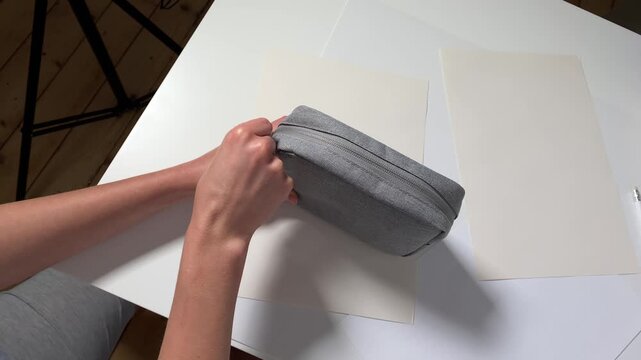 A close-up video shows a person's hands unzipping a grey fabric pouch or pencil case. The action takes place over a white desk with papers, shot from a high angle.