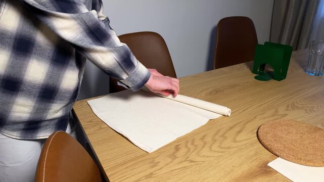 A person in a plaid shirt carefully rolls a white cloth placemat or napkin on a wooden dining table. The scene shows preparation for a meal or tidying up afterwards in a modern home setting.
