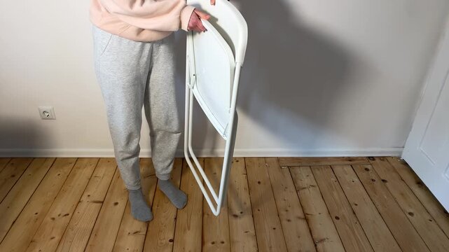 A person in casual wear unfolds a white folding chair inside a room with a wooden floor. This clip shows the setup process for temporary or event seating.