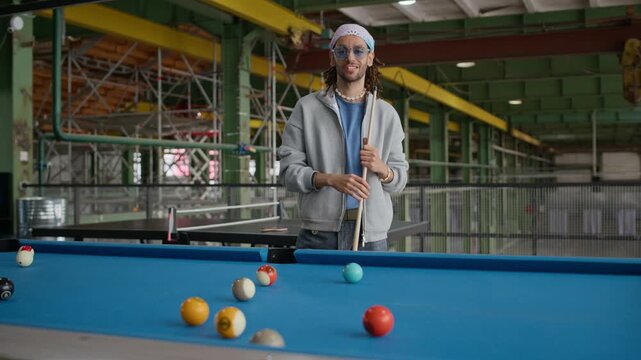 Medium portrait of young Biracial man in hipster outfit standing by pool table holding cue and looking at camera in loft interior