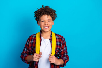 Smiling teenage boy with yellow backpack and plaid shirt standing against bright blue background, school student portrait © deagreez