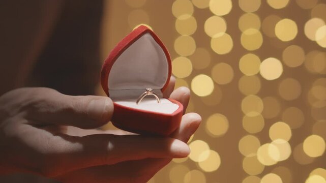 A closeup shows a hand holding an open red ring box containing a sparkling engagement ring, set against a soft bokeh backdrop that evokes romance and captures a moment of love and commitment