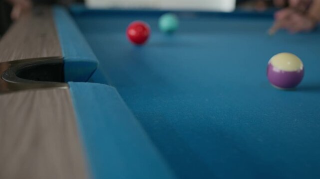 Extreme close up of pool ball falling into pocket on blue billiard table while unrecognizable player striking cue ball in blurred background, selective focus action shot