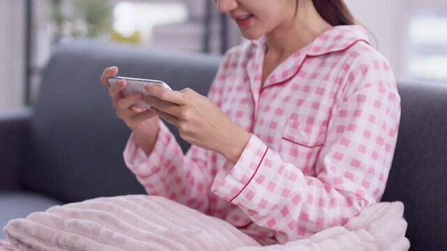 Asian adult woman wearing headphones plays mobile game on smartphone relaxing on grey sofa in modern home living room weekend leisure