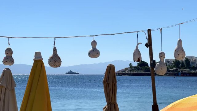 Traditional decorative gourd lamps hang above a sunny beach in Bitez, Bodrum. Folded sun umbrellas and a shimmering blue Mediterranean bay with distant yachts create a relaxing holiday scene.
