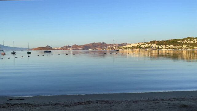 A tranquil sunrise over Bitez Bay, Bodrum. Sailboats rest on glassy, mirror-like water, reflecting the golden light and coastal hills of the Turkish Riviera in a serene, panoramic view.