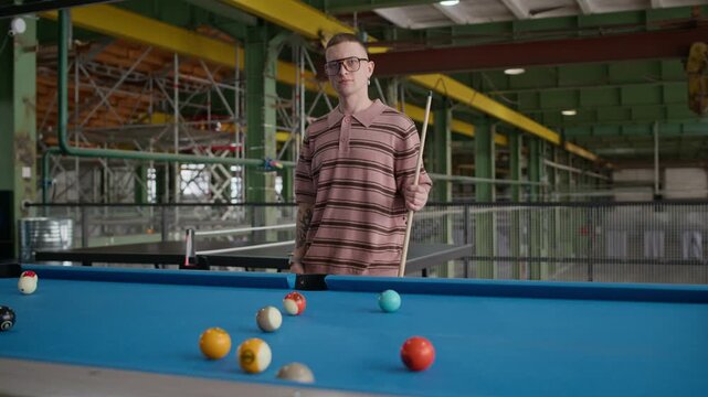 Medium portrait of young Caucasian man posing with billiard cue near blue pool table in industrial interior, showing confidence, style and leisure activity