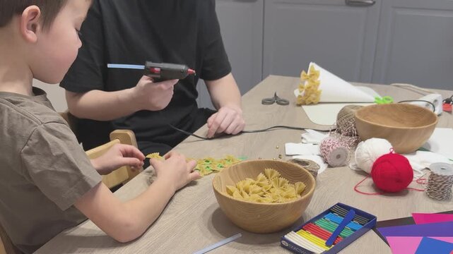 Mother and child work together using hot glue gun to attach pasta pieces to Christmas tree craft. Festive school or kindergarten project. Useful for parenting, holiday DIY, educational content and