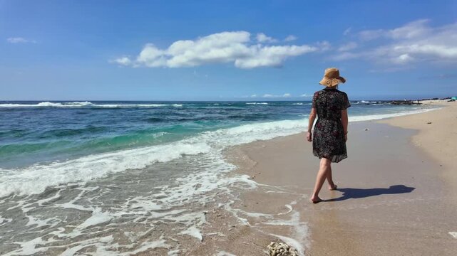 Traveler girl walking in a floral black dress on a tropical beach. Young girl traveler walking in nature. Traveler girl in a dress walking on the beach. Girl traveler walking on the beach. Summer trop