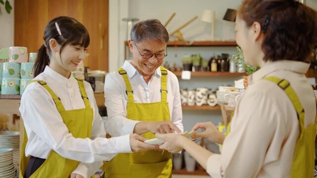 Three diverse shop employees in yellow aprons use a digital tablet to manage inventory and discuss products in a modern ceramic store. Professional teamwork in a cozy small business retail environment