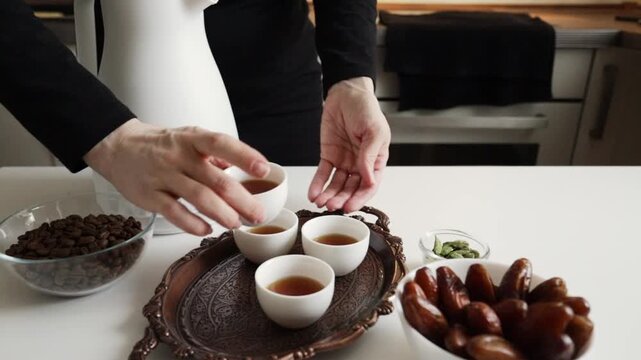 close-up of hands placing a filled white finjan cup onto a copper tray with arabic coffee, coffee beans, dates and cardamom prepared for serving