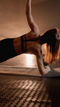 Young fit woman holds plank pose on mat in modern fitness studio, illuminated by warm sunlight. Intense workout atmosphere highlights dedication and healthy lifestyle