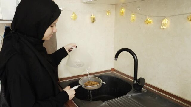 muslim woman in black hijab rinses golden raisins over the sink in a home kitchen decorated with warm festive lights, documenting an ingredient-prep moment for a traditional meal